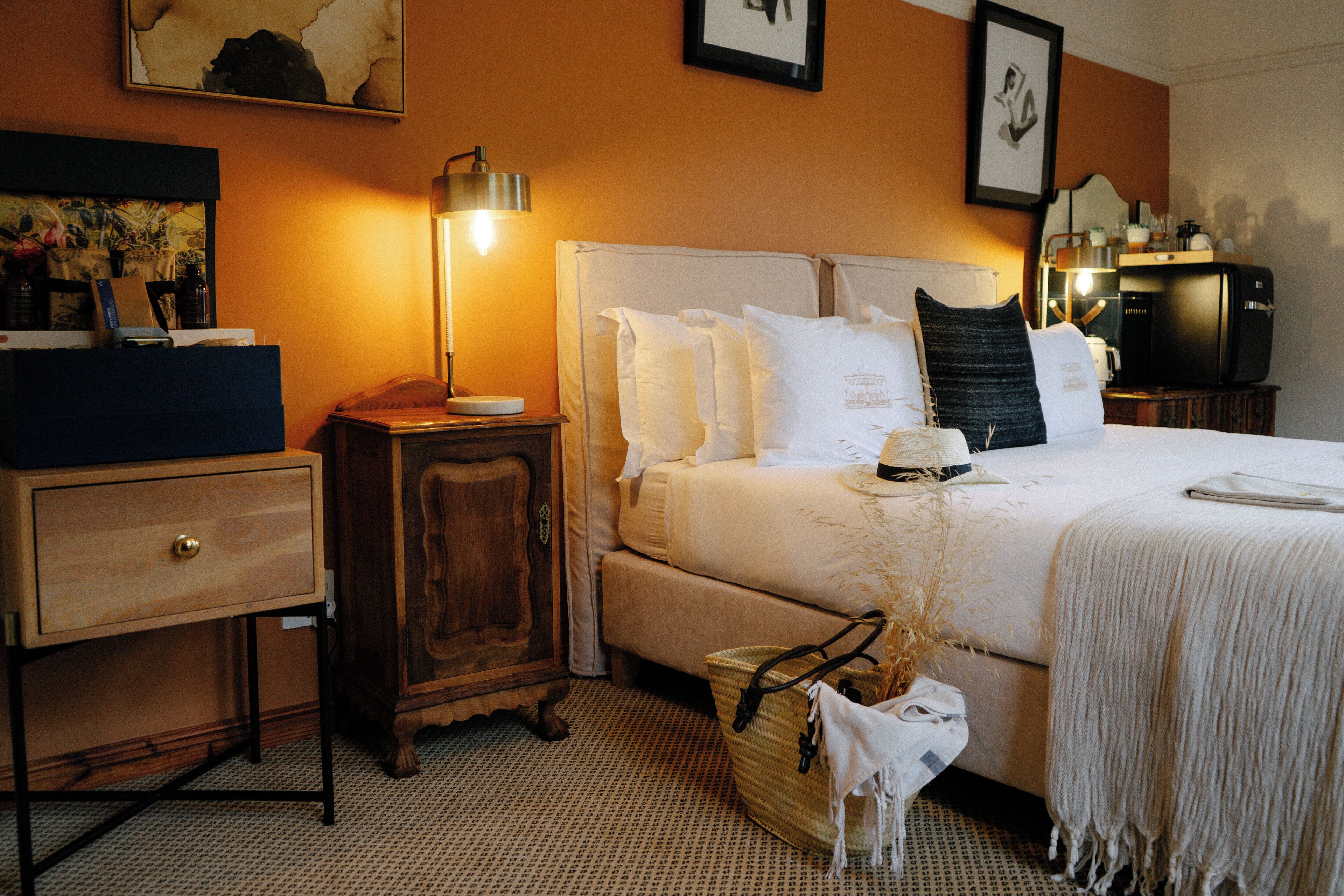 A hotel room with a bed, bedside table, lamp, dresser, and a basket with a towel.