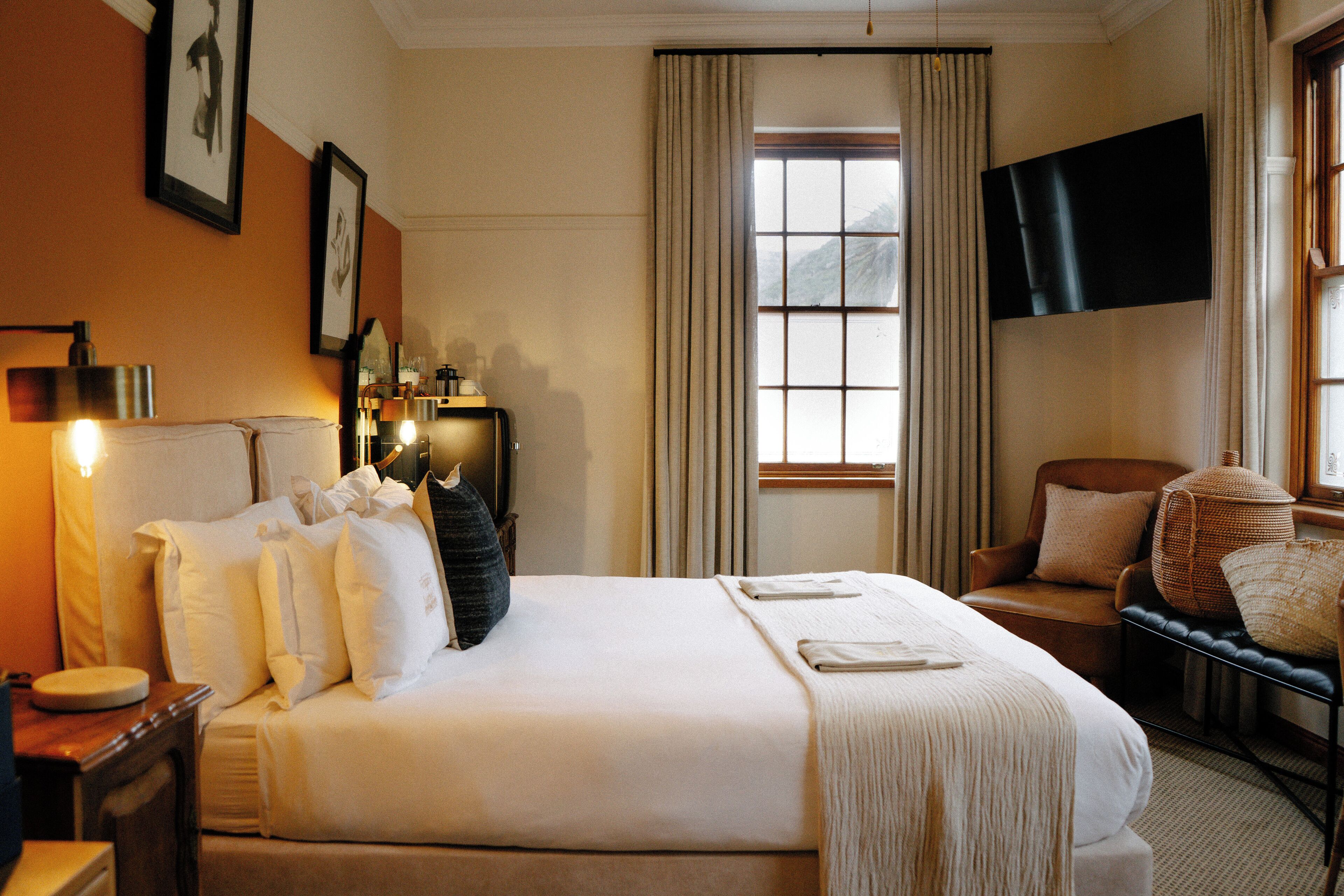A hotel room with a large bed, a sitting area with a chair and a small table, a TV mounted on the wall, and a window with curtains.