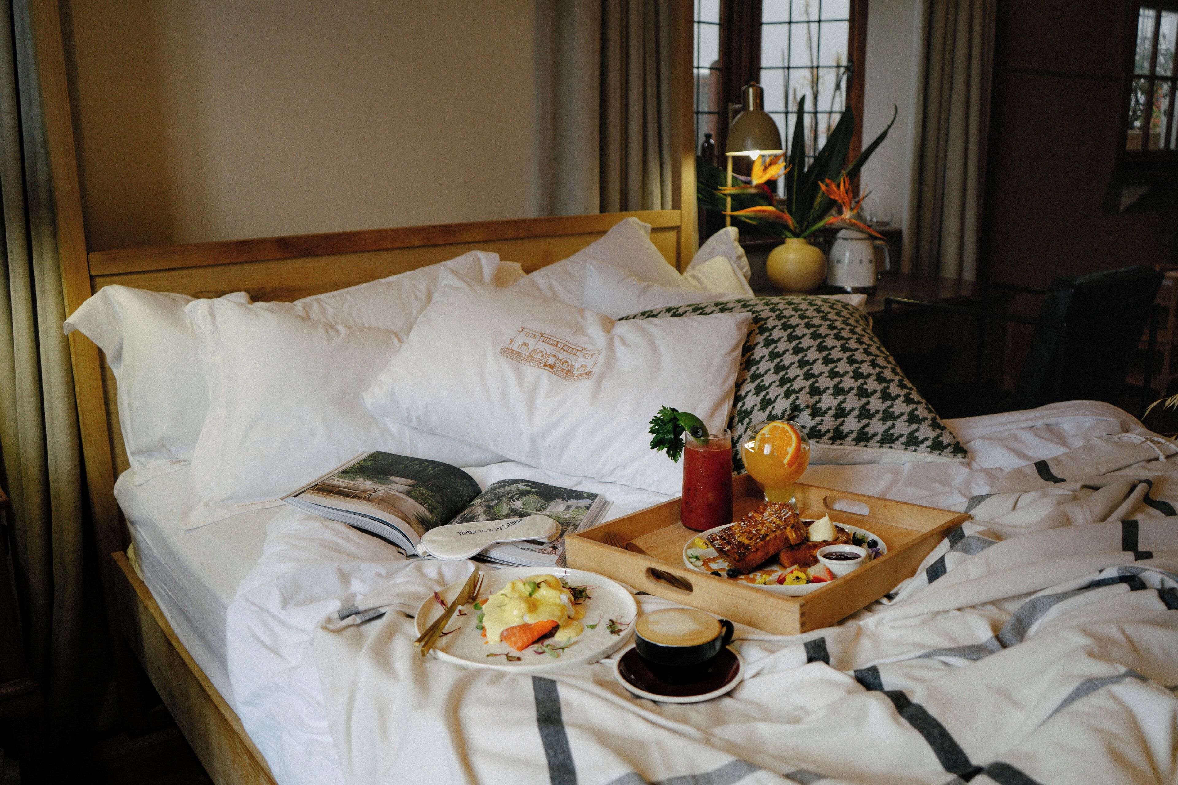 A neatly made bed with a tray of breakfast including eggs, toast, and juice, and a cup of coffee on a hotel room bed.