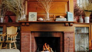 Een gezellige woonkamer met een haard van baksteen, een houten stoel en een boekenkast vol boeken en decoratieve objecten.