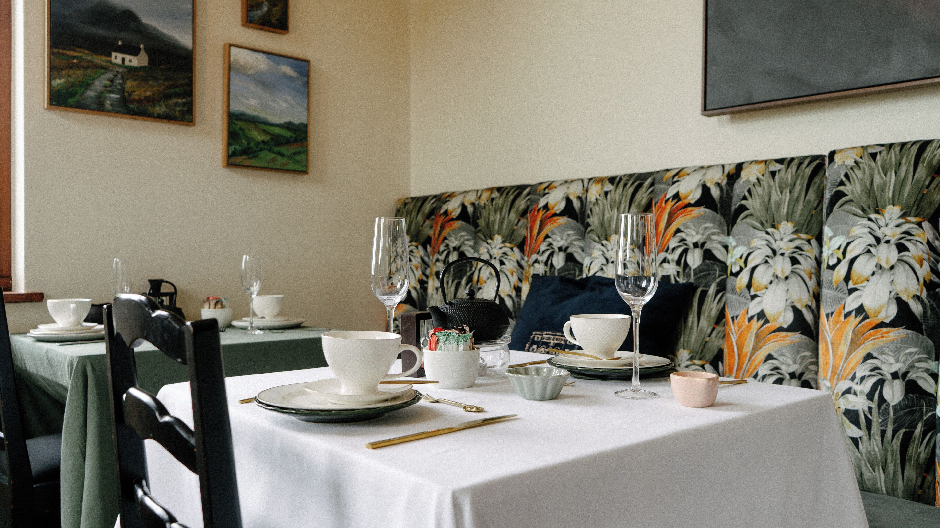 Une table à manger dressée avec des assiettes, des bols, des couverts et des verres, le tout sur fond d’un papier peint à motifs floraux et agrémentée d’œuvres d’art encadrées accrochées au mur.