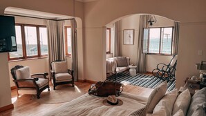 Een knus hotelkamer met een groot bed, een zithoek met een stoel en een klein tafeltje, uitzicht op de zee door de ramen, en een plafondventilator.
