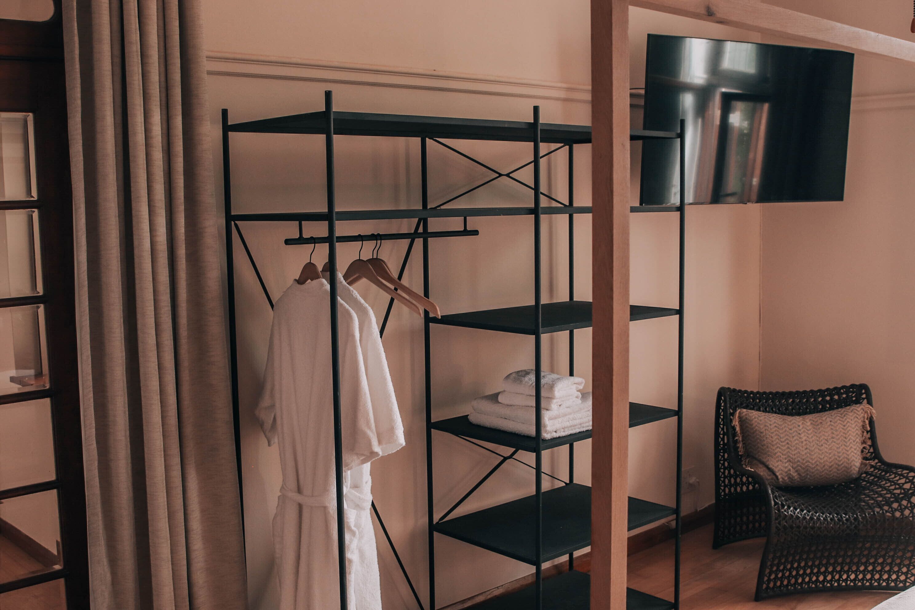 A room with a metal shelving unit holding towels, a white bathrobe, and a wicker chair with a cushion.