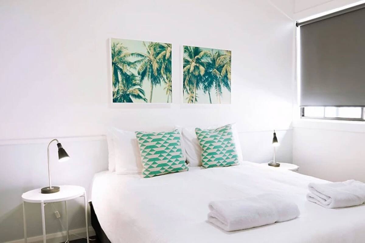 A hotel room with a large bed, two nightstands, a window with blinds, and two wall-mounted paintings of palm trees.