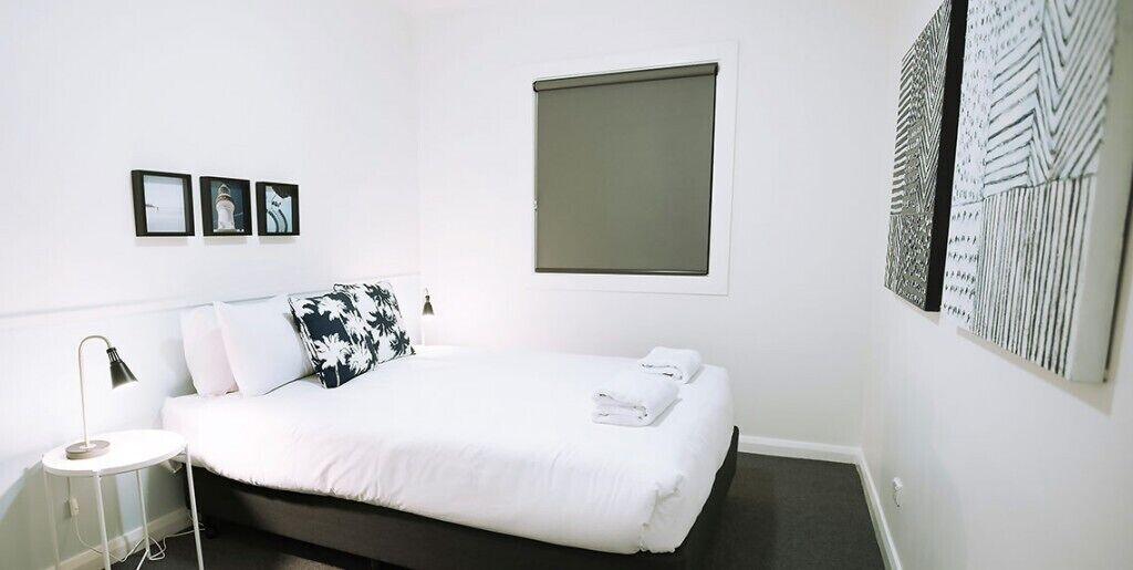 Ein modernes Hotelzimmer mit einem ordentlich bezogenen Bett, einem Nachttisch mit Lampe und einem großen Fenster mit Blick auf die Stadt.