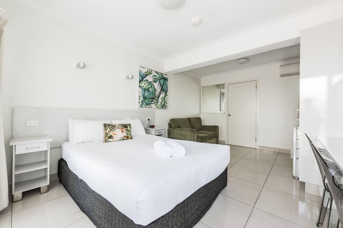 A modern hotel room with a large bed, a nightstand, a green armchair, and a desk.