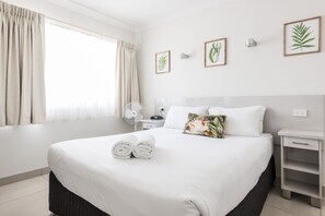 A neatly made bed with white linens and two rolled towels, a bedside table with a lamp, and a wall-mounted fan. - Hibiscus Motel (Byron Bay)