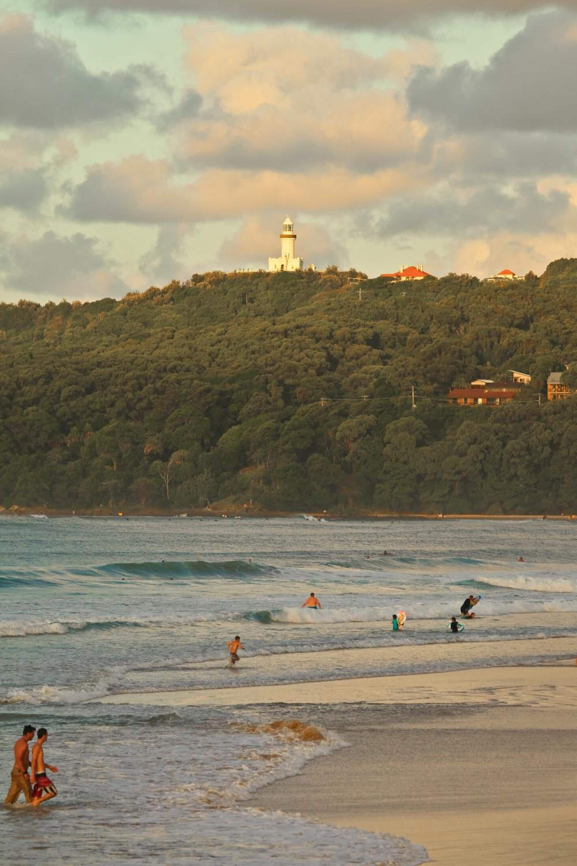 A beach with people swimming and walking, a lighthouse in the background, and a hill with trees and buildings.