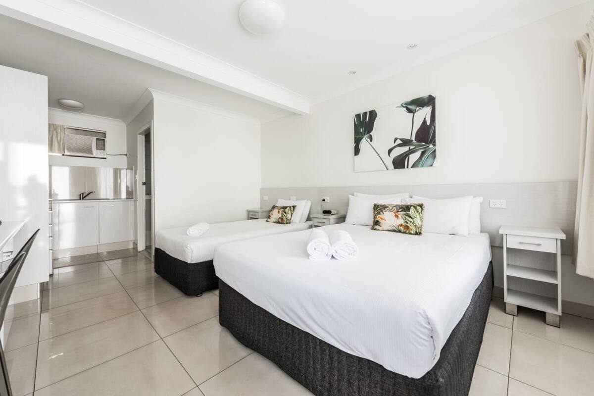 A modern hotel room with a large bed, two bedside tables, a small desk, and a kitchenette.
