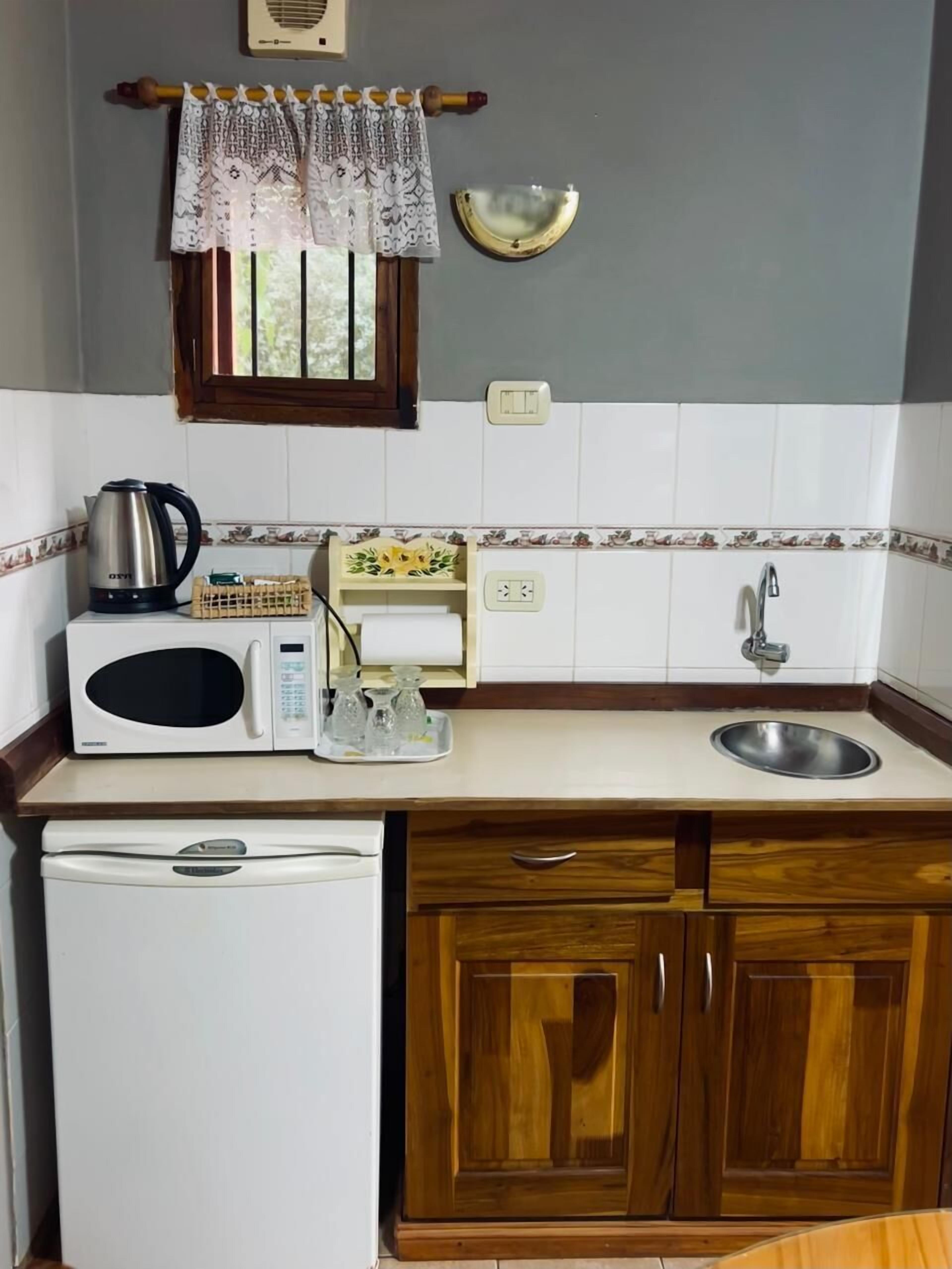 Une cuisine compacte équipée d’un four à micro-ondes, d’une bouilloire et d’un petit réfrigérateur. On y trouve un meuble en bois avec un évier et une fenêtre au-dessus du plan de travail.