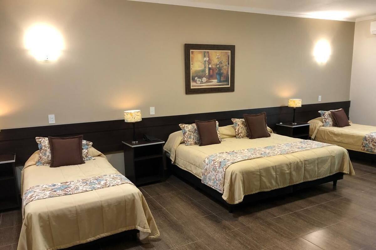 Ein Hotelzimmer mit drei Betten, jedes mit einer blumigen Tagesdecke und braunen Kissen. An der Wand über den Betten hängt ein Gemälde und zwischen den Betten befindet sich ein Nachttisch mit einer Lampe.