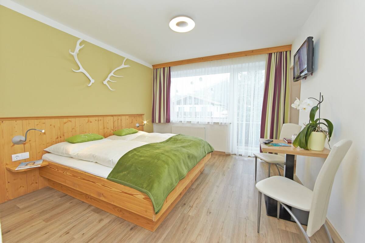 Ein modernes Hotelzimmer mit einem hölzernen Bett, einem Schreibtisch, einem Sessel, einem Fernseher und einem Fenster mit Vorhängen.