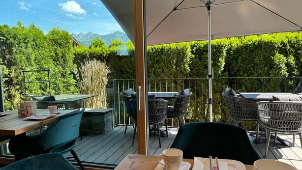 An outdoor dining area with tables and chairs, a large umbrella, and a view of green trees and mountains.