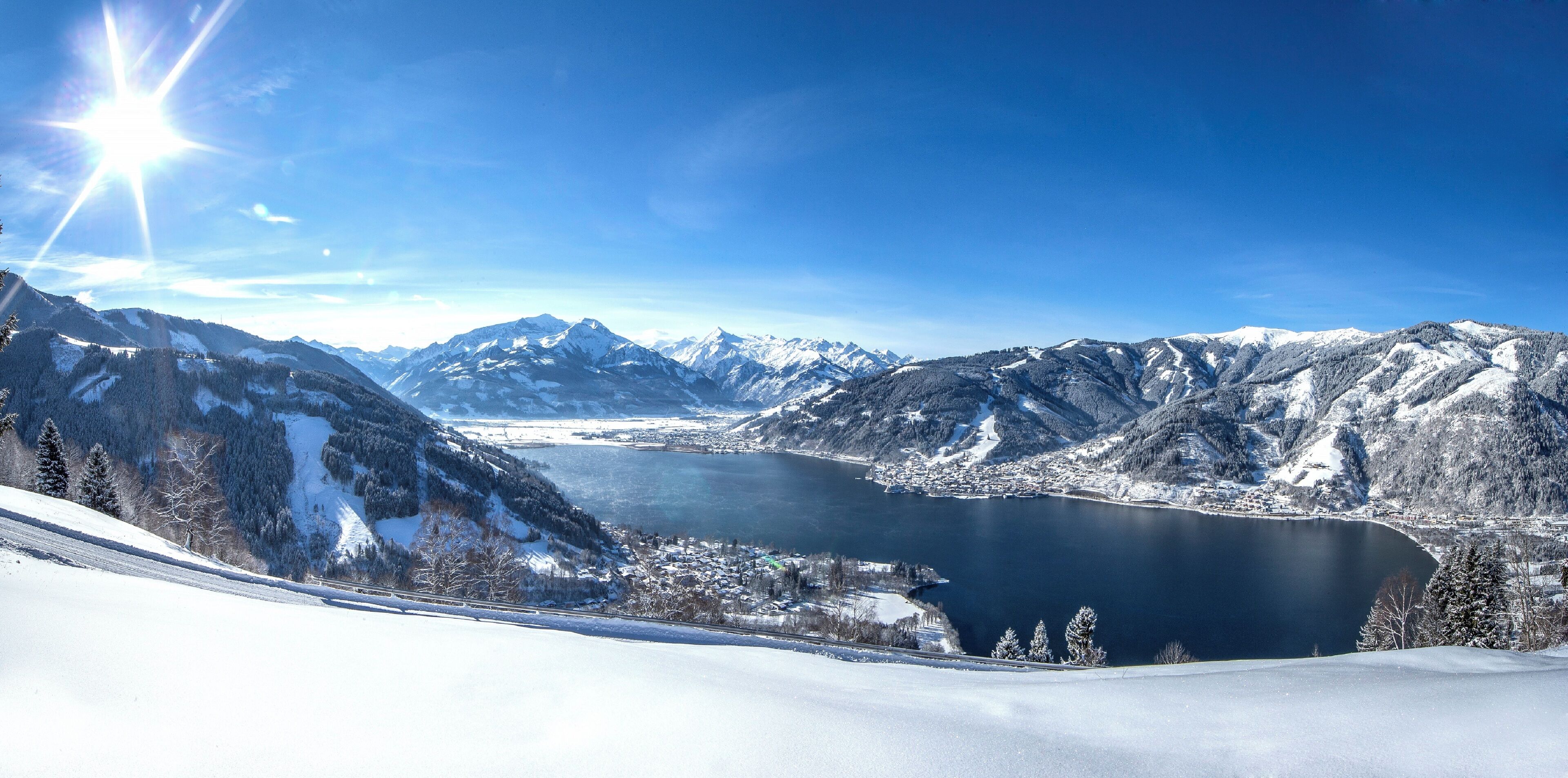 A panoramic view of a mountainous landscape with a lake, snow-covered peaks, and a clear sky.