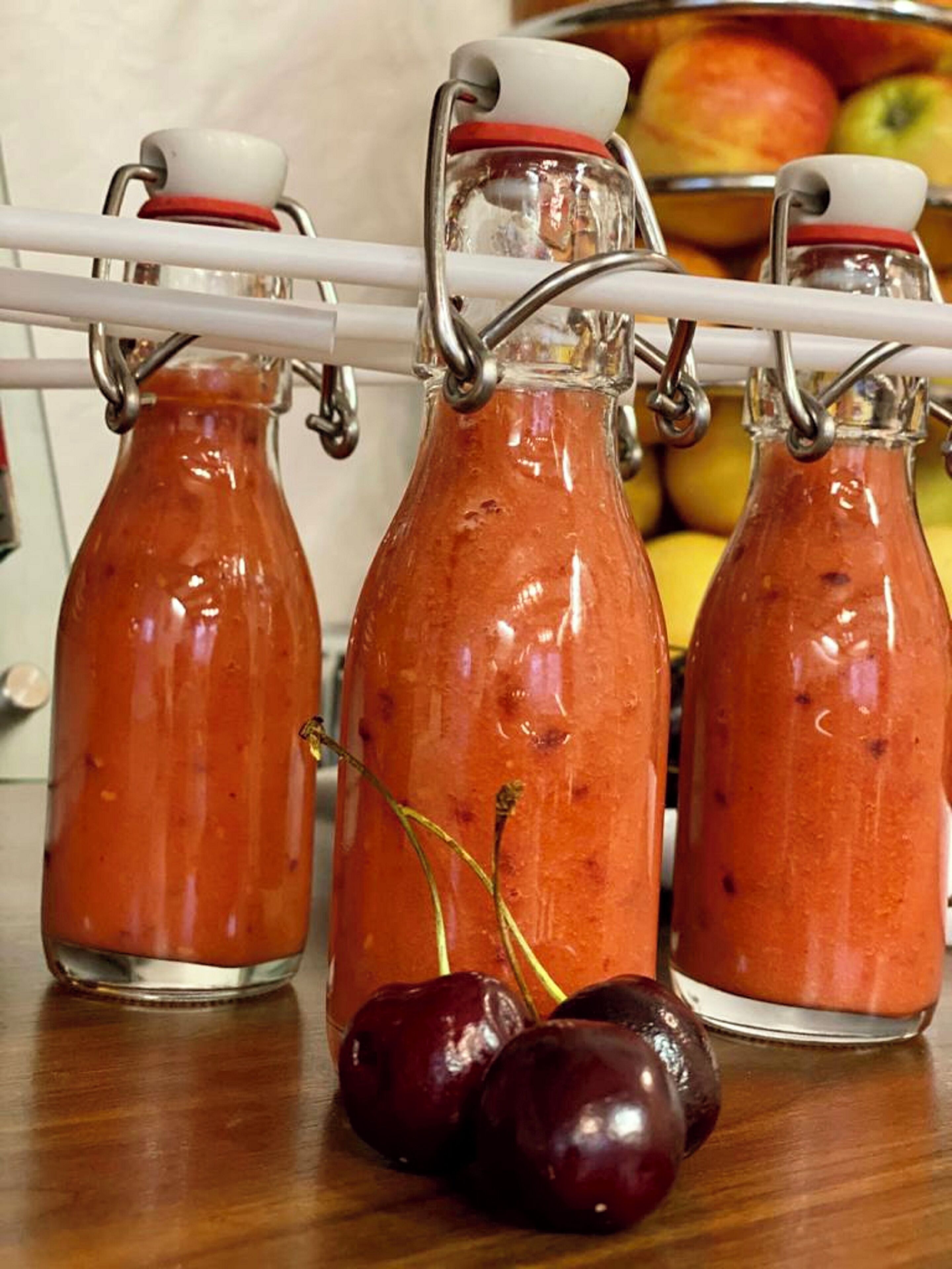 Trois bouteilles en verre munies de fermoirs métalliques, contenant chacune un liquide rouge, vraisemblablement une sauce ou un condiment, avec une cerise placée devant la bouteille du milieu.