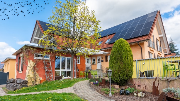 Una casa de dos pisos con techo inclinado, paneles solares, un jardín con flores y un camino pavimentado.