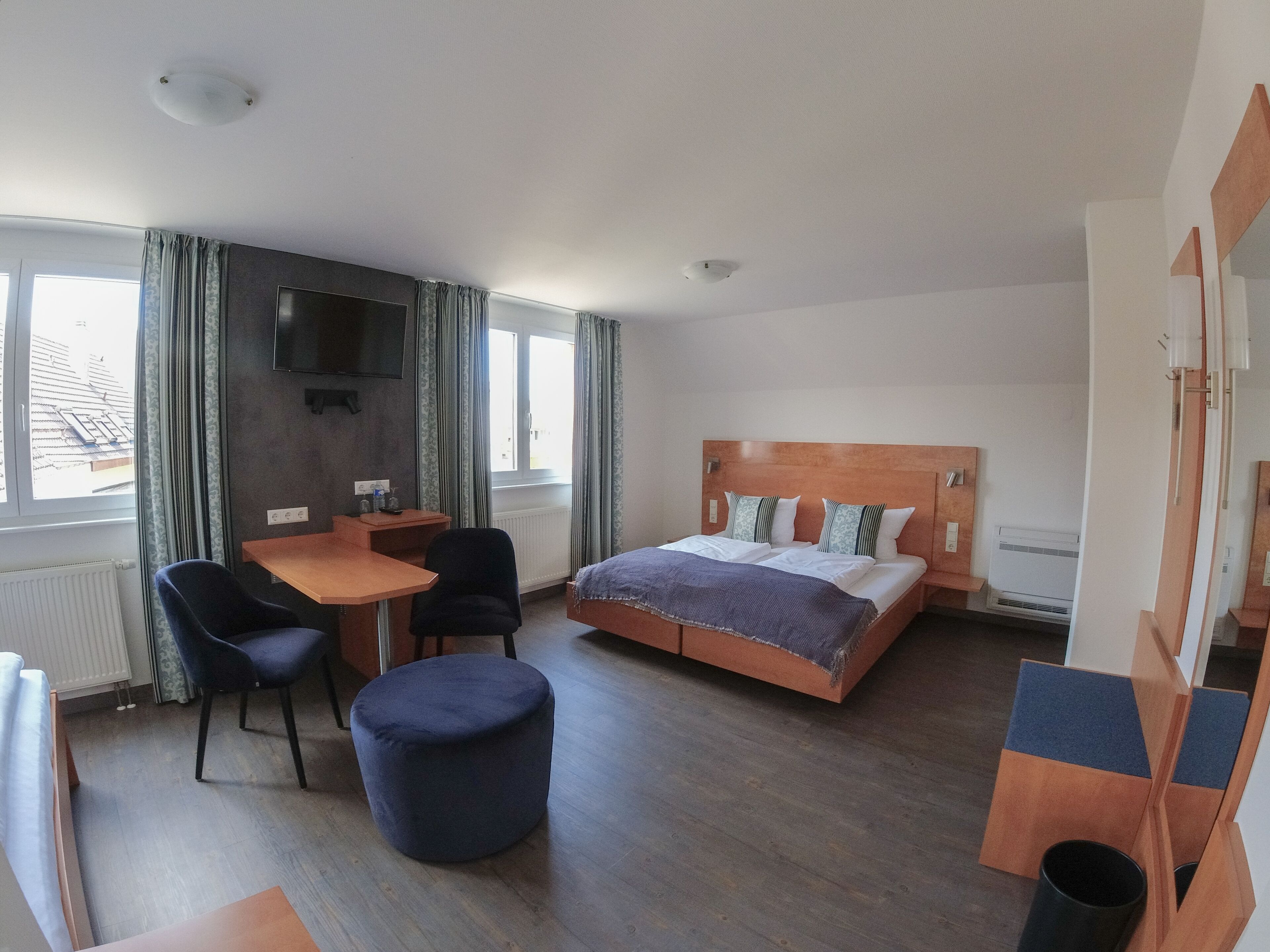 A hotel room with a bed, a desk, a chair, a round ottoman, and a TV mounted on the wall.