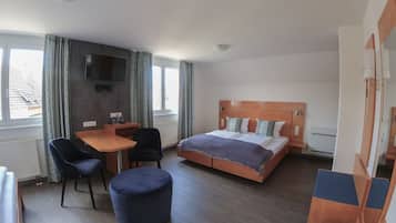 A hotel room with a bed, a desk, a chair, a round ottoman, and a TV mounted on the wall.