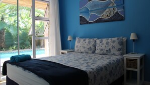 Un dormitorio con una cama, dos mesitas de noche con lámparas, un cuadro en la pared y vistas a una piscina y vegetación.
