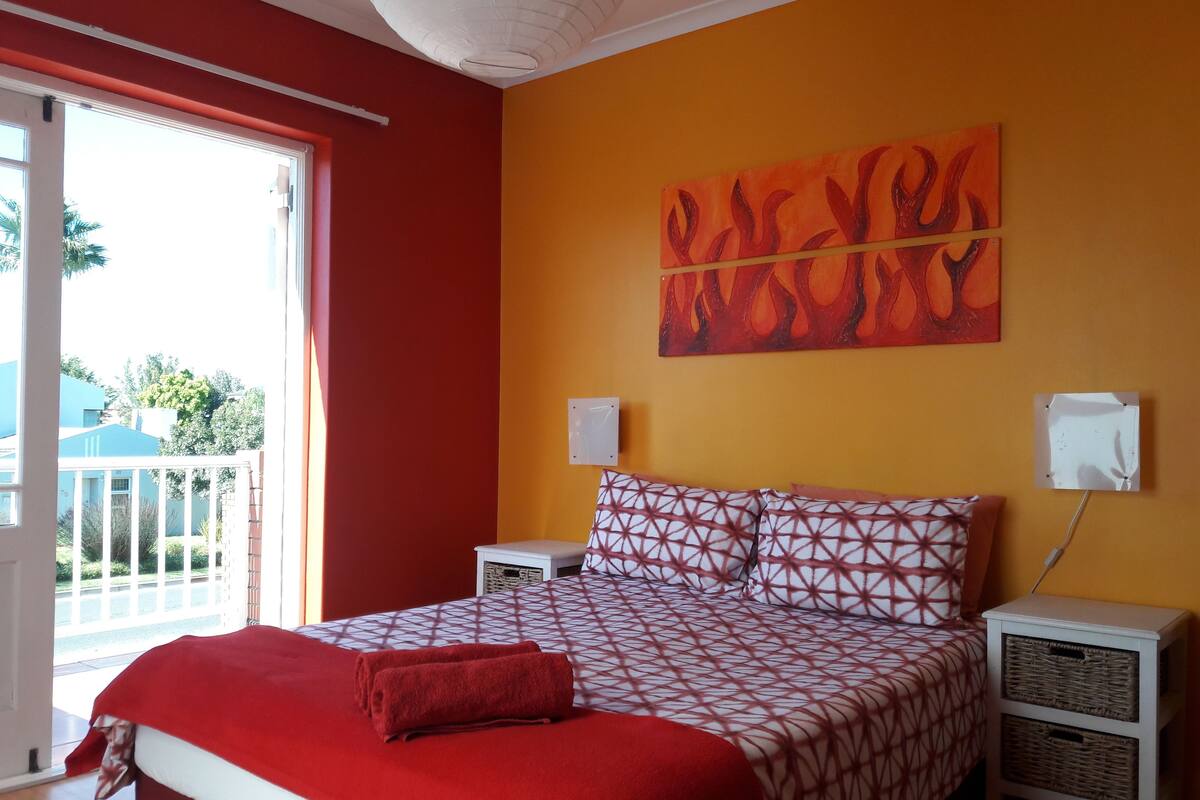 Un dormitorio con colcha y funda roja y blanca, una manta roja y un cojín estampado. Hay dos mesitas de noche con cestas. Un cuadro grande de llamas adorna la pared.