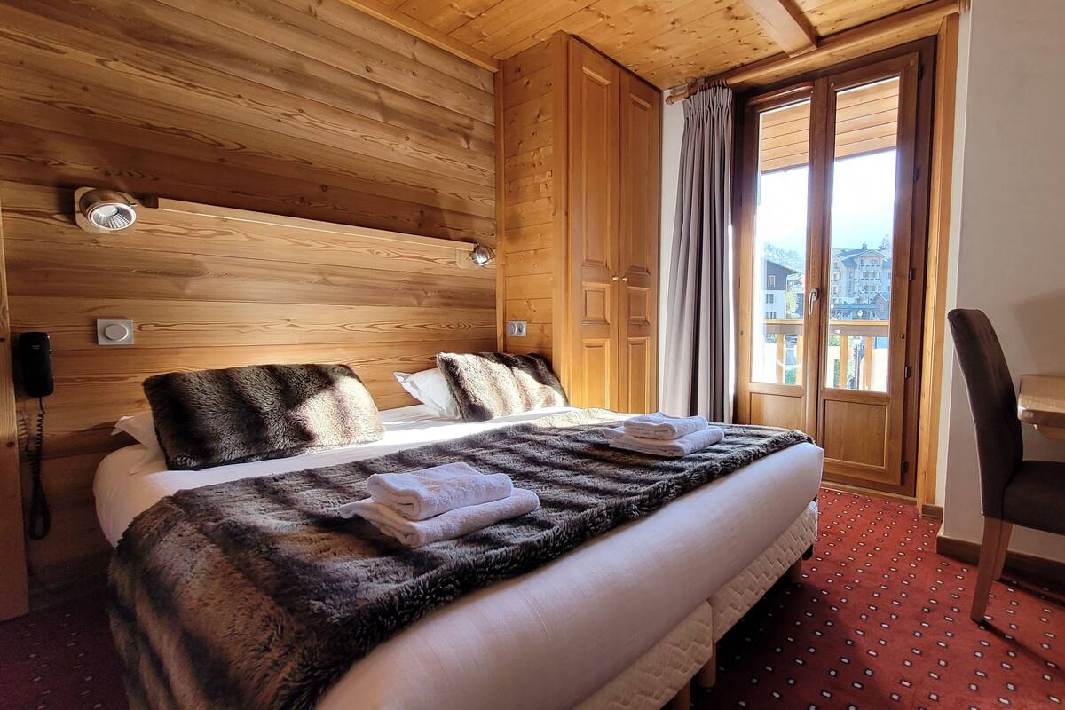 A wooden-paneled room with a bed, a desk, and a view of buildings through a door.