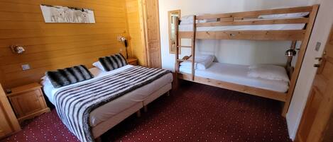 Una habitación con una cama litera, una cama individual, paredes de madera, una alfombra roja estampada, una mesita de noche pequeña y una lámpara fijada a la pared.