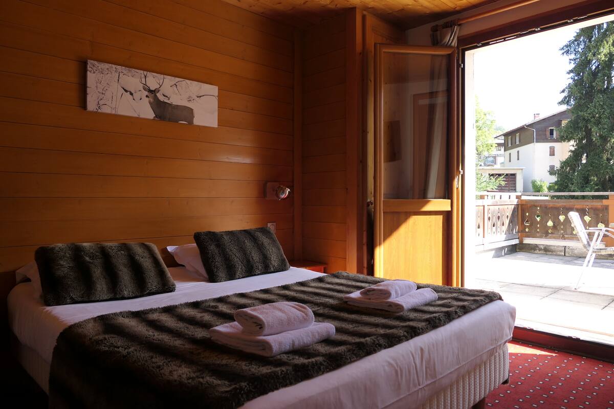 A wooden-paneled room with a bed, a painting, and a view of the outside.