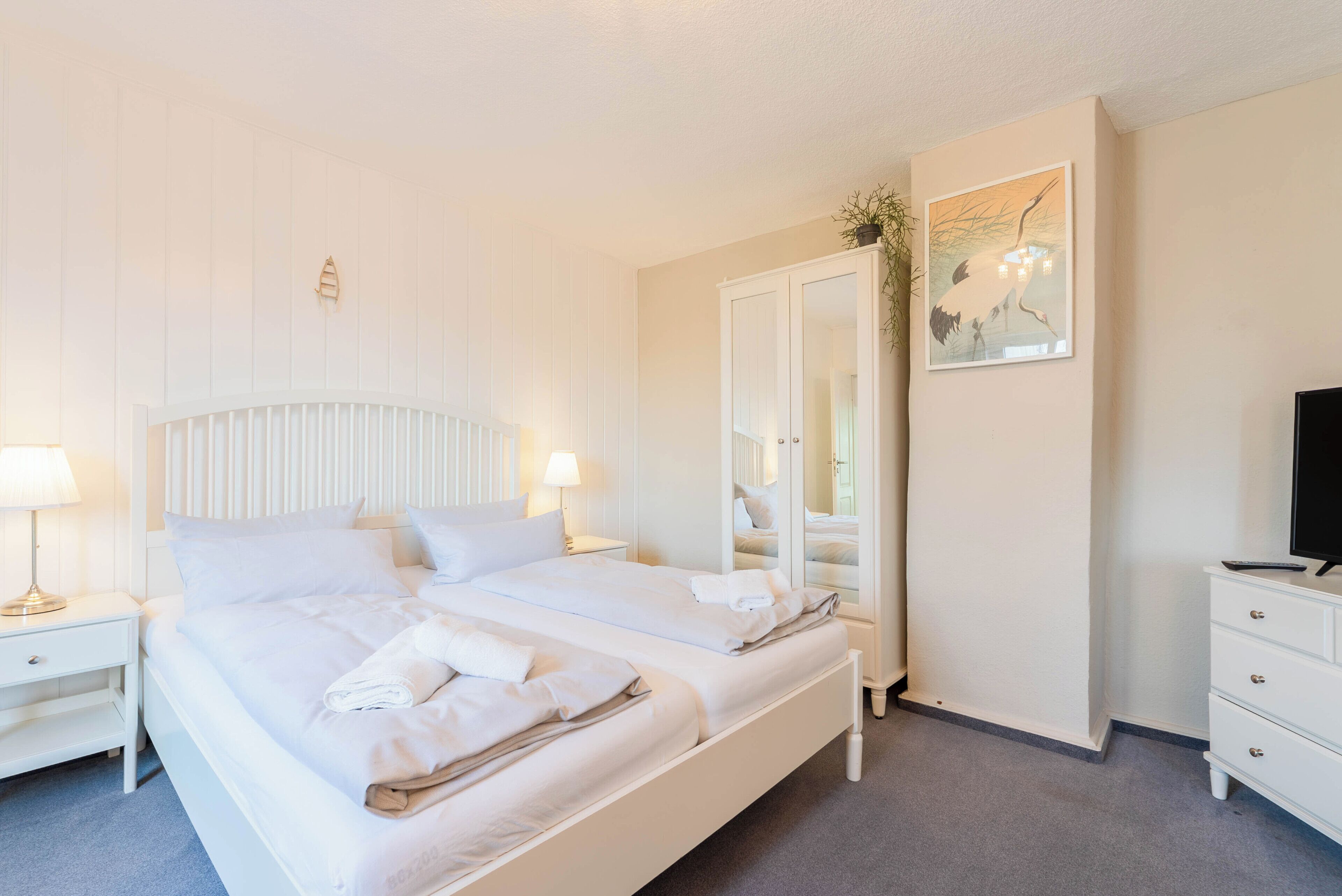 A double bed with white bedding, a nightstand with a lamp, a dresser, and a mirror.