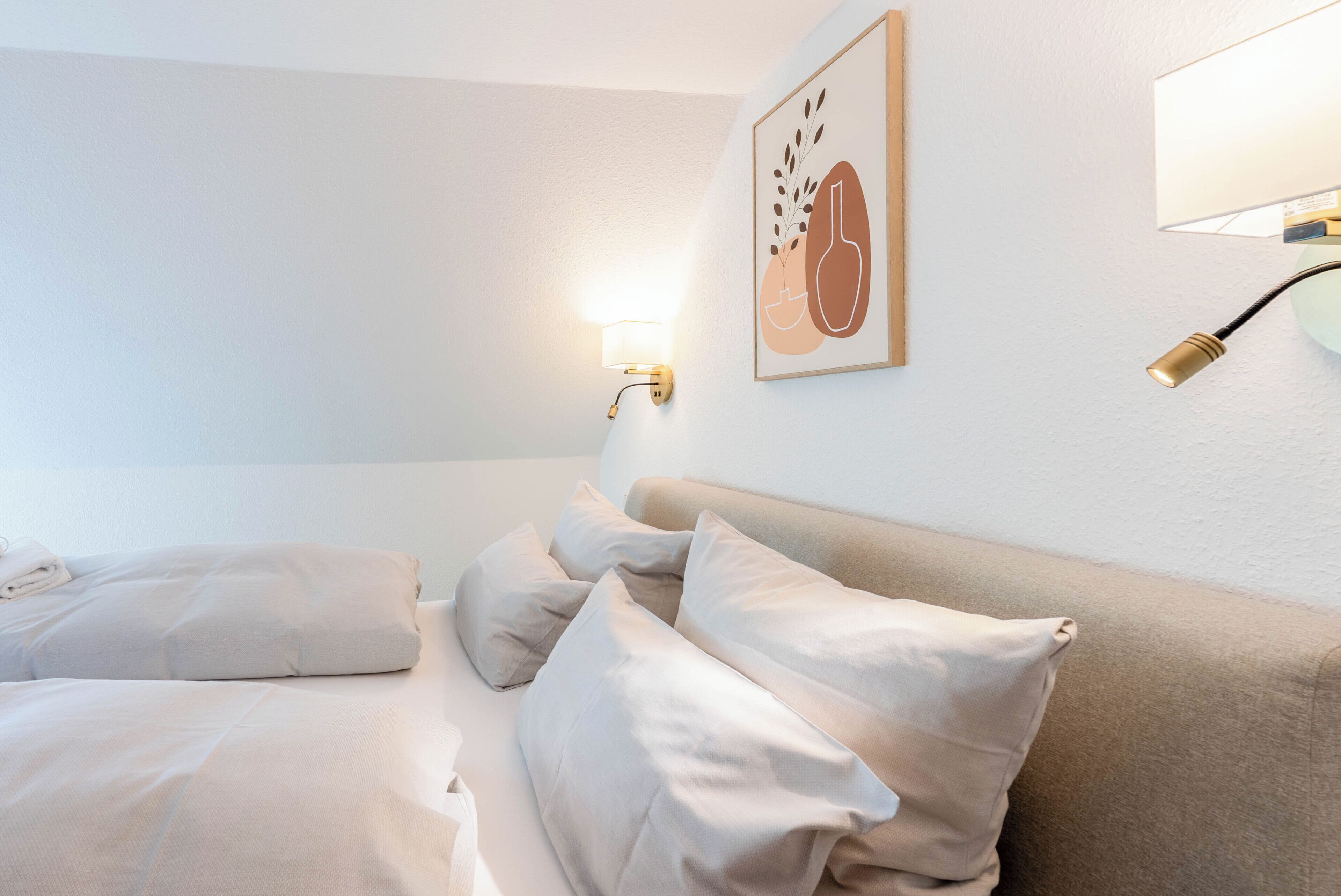 A hotel room with a bed, pillows, a wall-mounted light, and a framed picture.