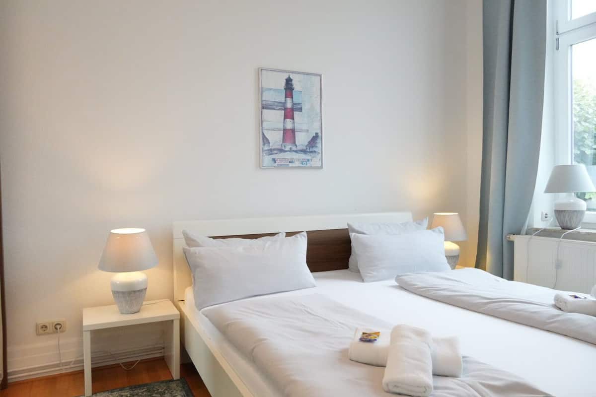 A hotel room with a bed, two bedside lamps, a nightstand, a painting of a lighthouse, and a window with curtains.