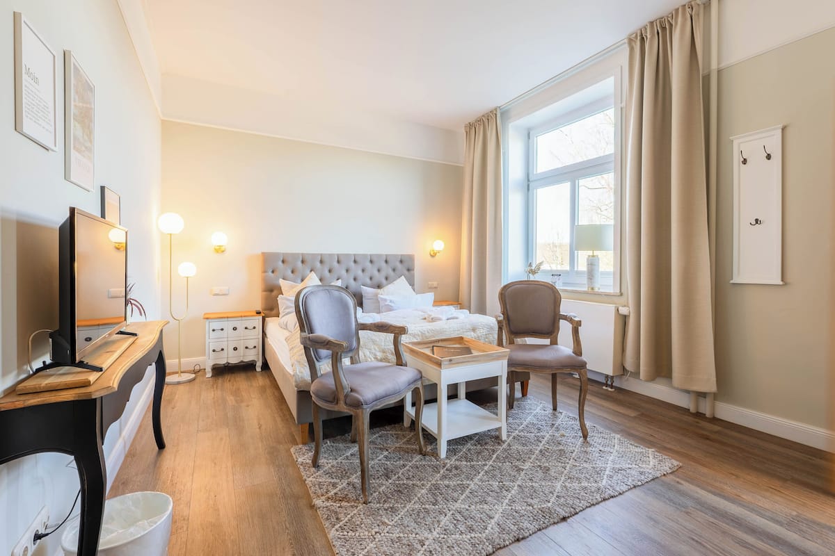 A hotel room with a bed, bedside tables, a desk, a chair, a lamp, and a window with curtains.