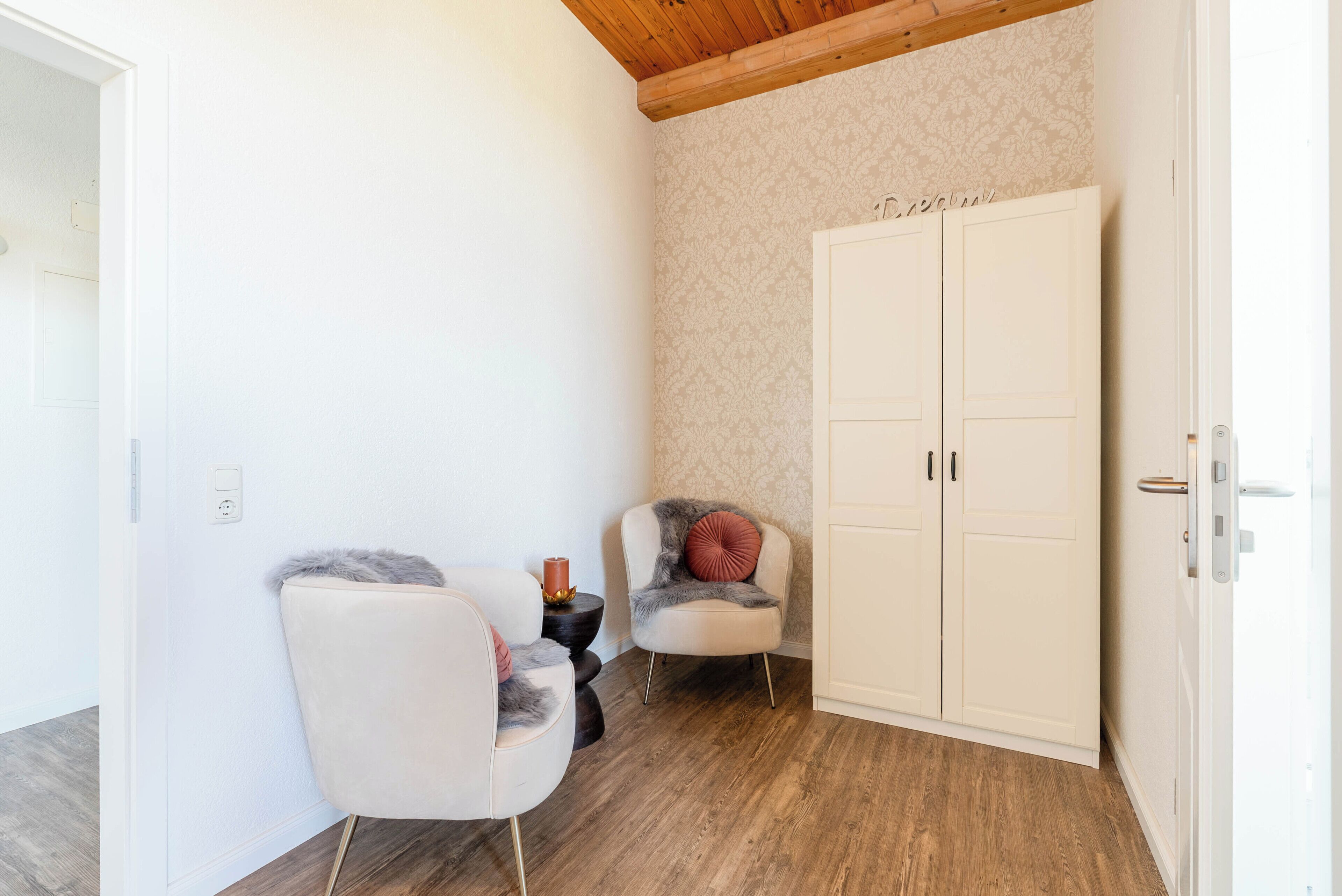 Ein Zimmer mit Holzbalkendecke, einem weißen Kleiderschrank, zwei Stühlen mit Kissen, einem kleinen Tisch mit Kerze und einer Wand mit Blumenmuster.