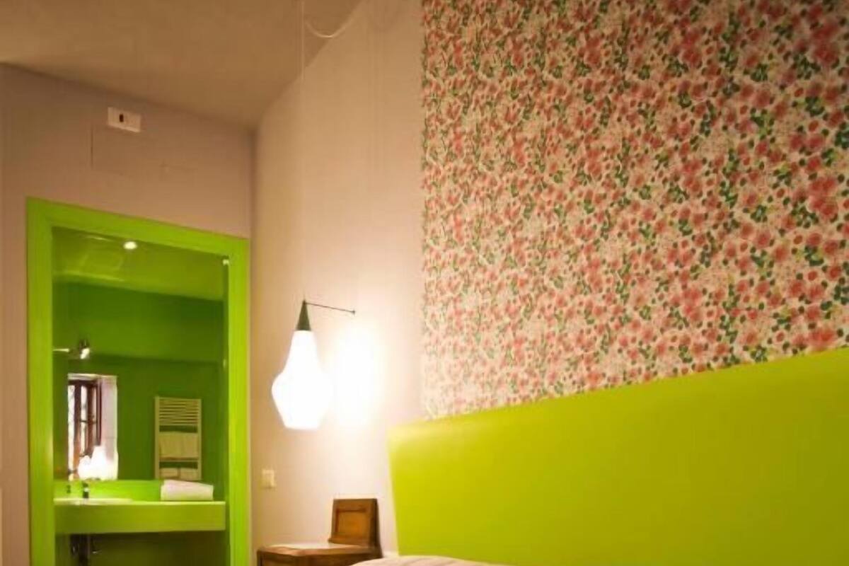 A room with a green door leading to a bathroom, a bed with a quilted cover, and a wall with a floral pattern.