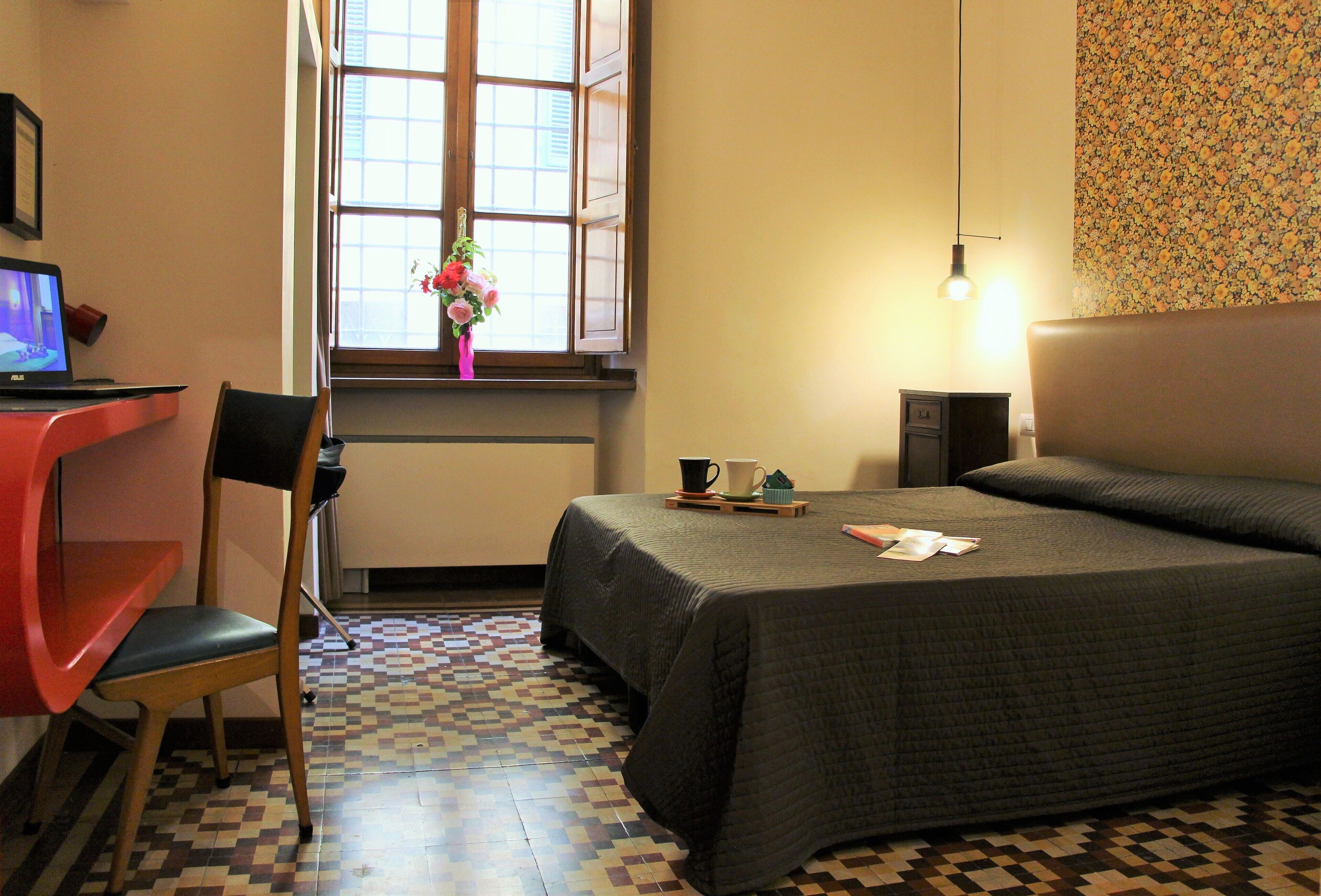 A hotel room with a bed, a desk with a laptop, a chair, a window with a view, and a patterned floor.