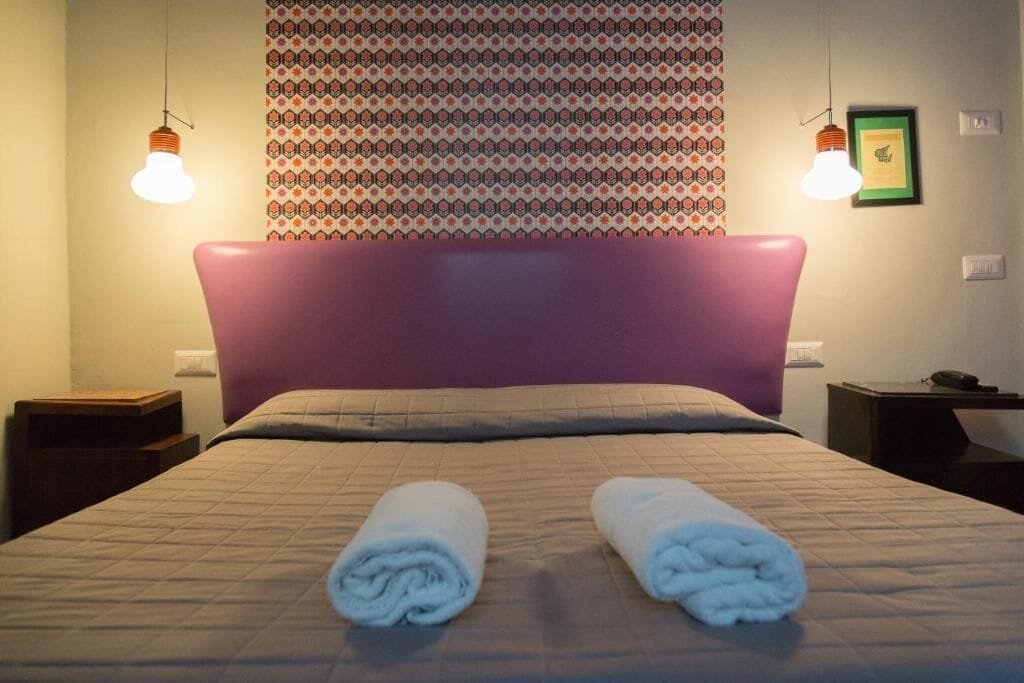 Una cama bien hecha con cabecera, dos toallas enrolladas y un tapiz decorativo en la pared.