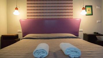 Una cama bien hecha con cabecera, dos toallas enrolladas y un tapiz decorativo en la pared.