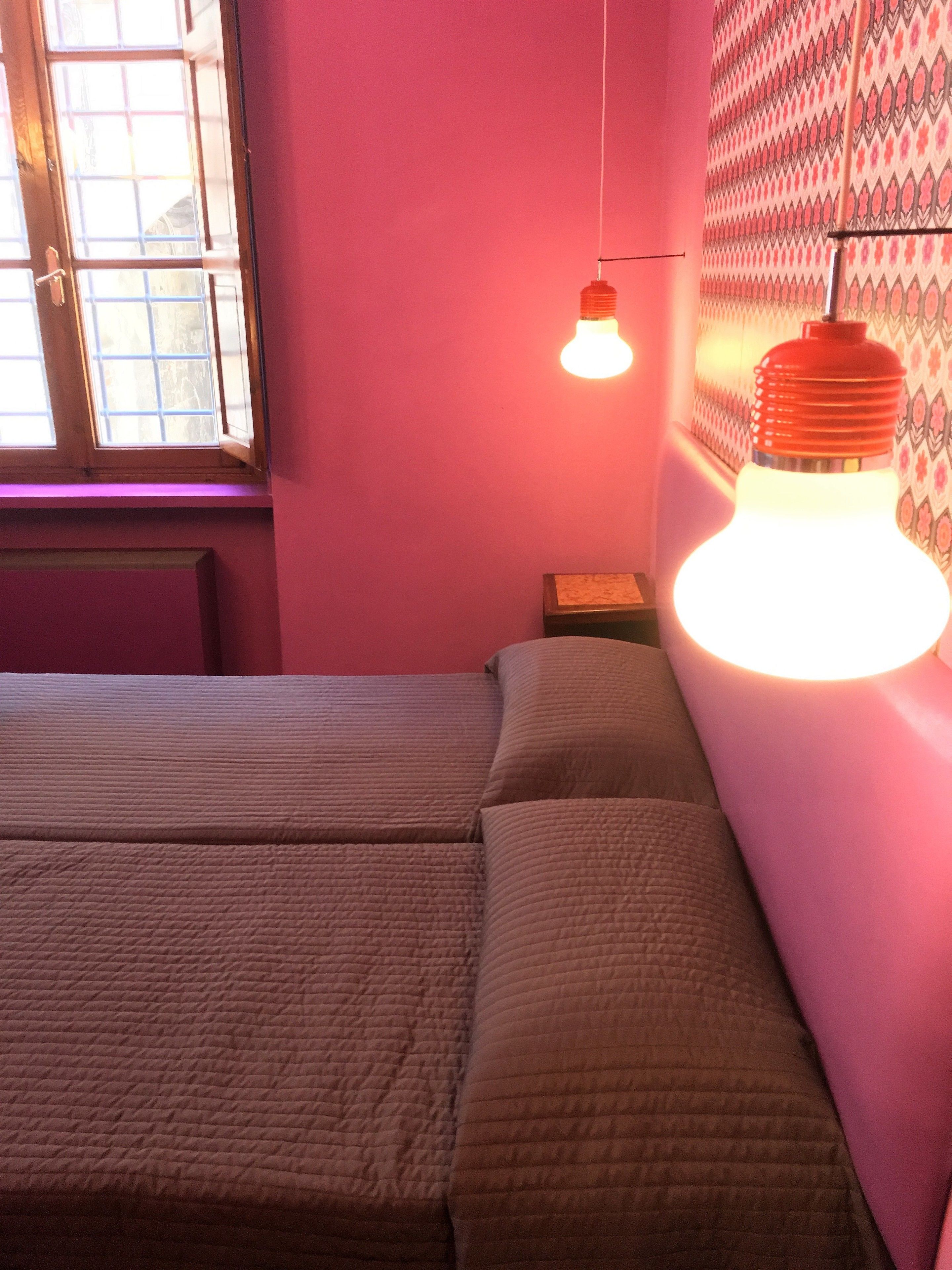 A room with a bed, a nightstand, a wall-mounted light fixture, and a window.