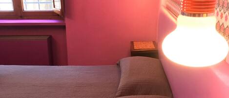 A room with a bed, a nightstand, a wall-mounted light fixture, and a window.