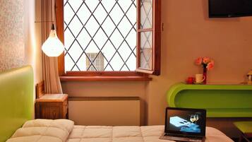 Una habitación de hotel con una cama, un ordenador portátil sobre la cama, una ventana con vistas, un banco verde y una mesita con una taza y flores.