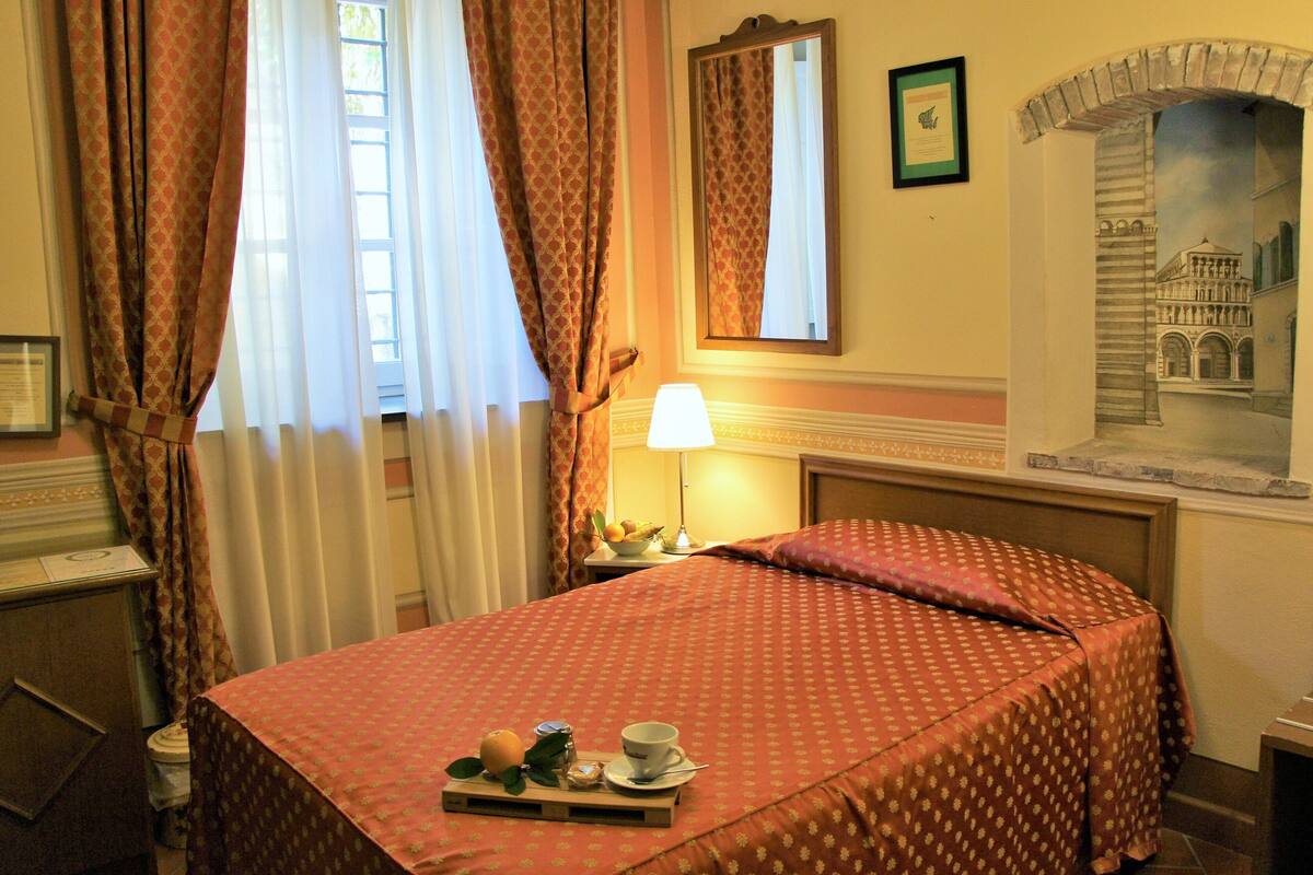 A hotel room with a bed, a nightstand with a lamp, a tray with breakfast items, and a painting of a historical building.