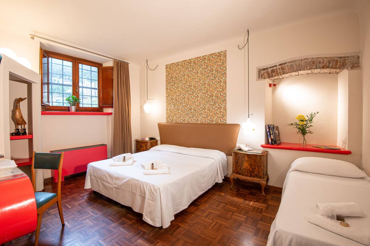 A hotel room with a large bed, a desk with a chair, a window with curtains, a wall with a floral pattern, and a small table with a vase.