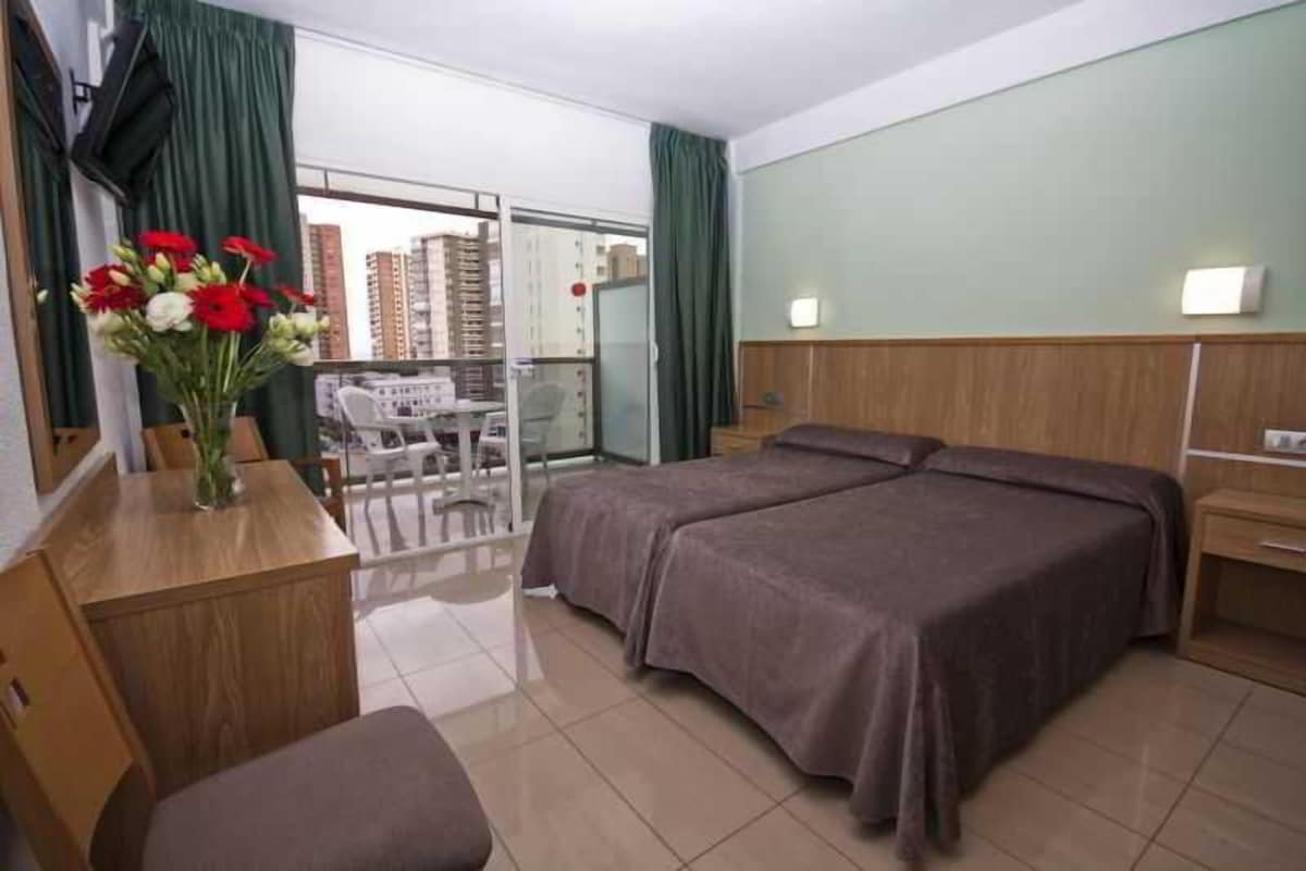 A hotel room with a bed, a desk, a chair, and a vase of flowers.