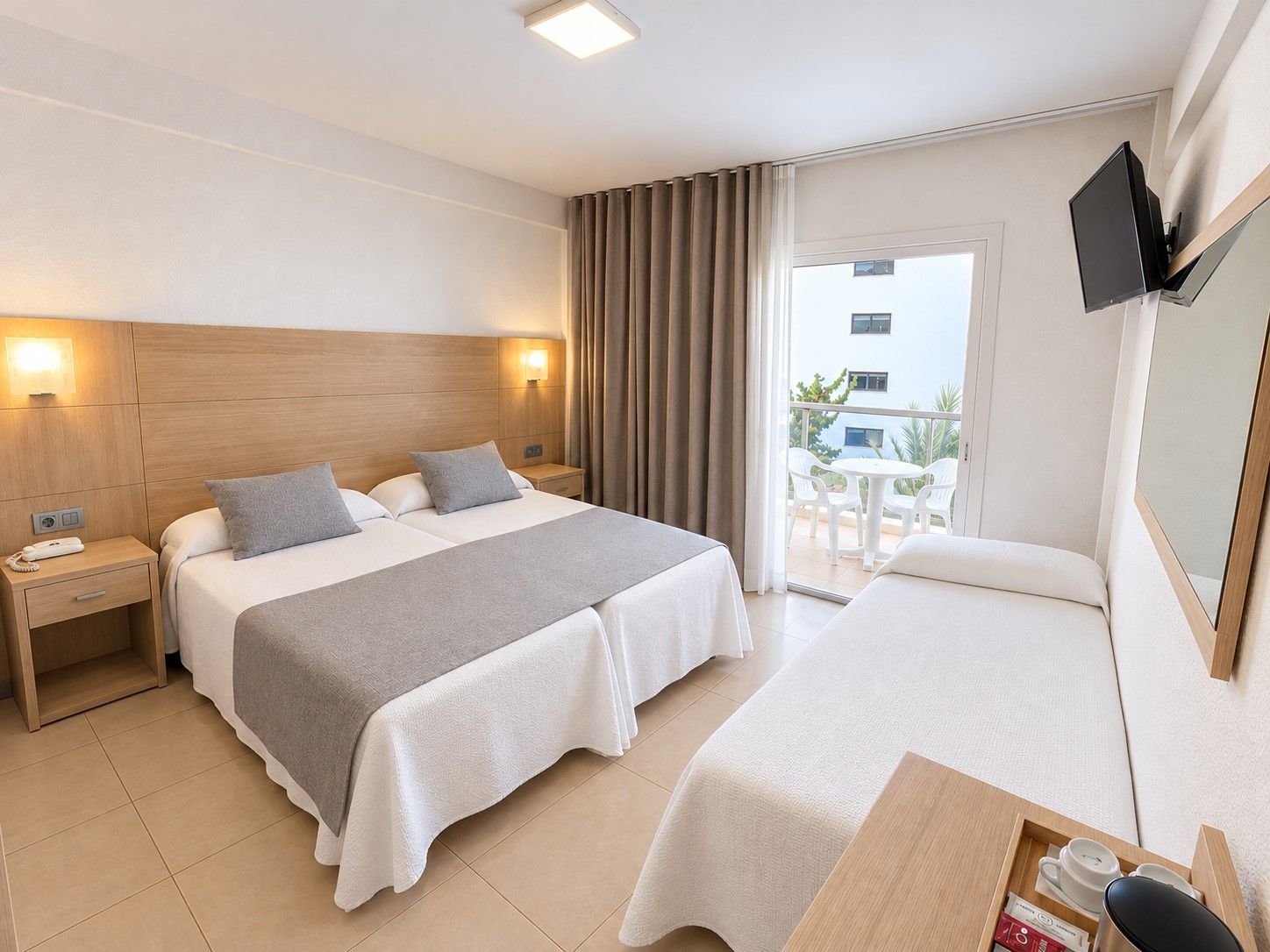 Habitació d'hotel amb un llit gran, una taula de nit amb una làmpada, un televisor fixat a la paret i un balcó amb mobles d'exterior.