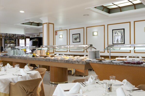 A buffet setup with multiple serving stations, a bar area with bottles, and neatly arranged tables with white tablecloths and chairs.