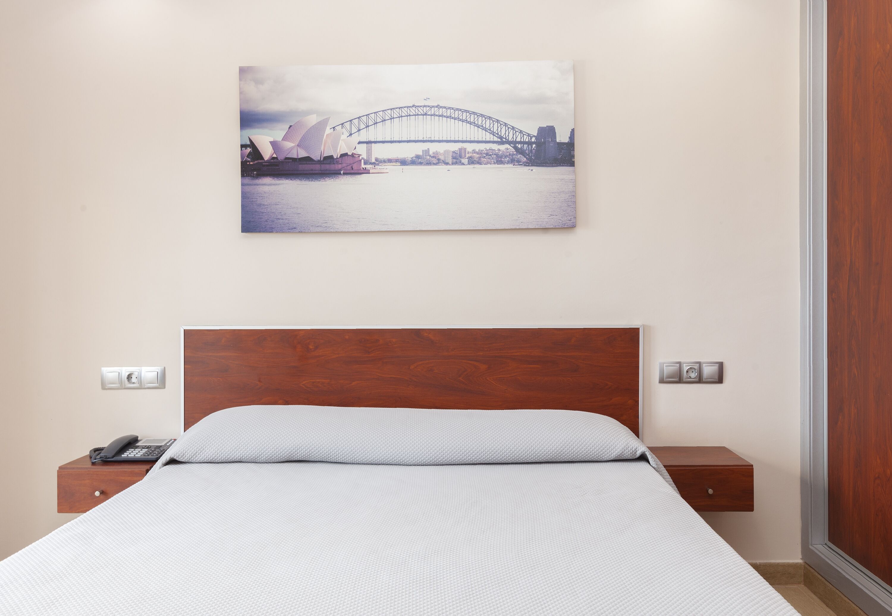 Habitación de hotel con una cama, cabecera de madera, mesita de noche con teléfono y un cuadro colgado en la pared de un puente.