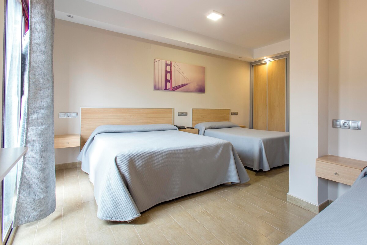 Une chambre d’hôtel avec deux lits, une tête de lit en bois, une table de chevet, une commode, un tableau et une fenêtre avec un rideau.
