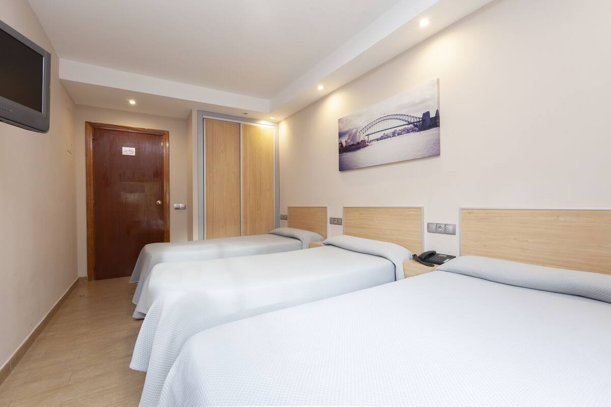 A hotel room with three single beds, a wooden door, a painting of a bridge, and a mounted television.