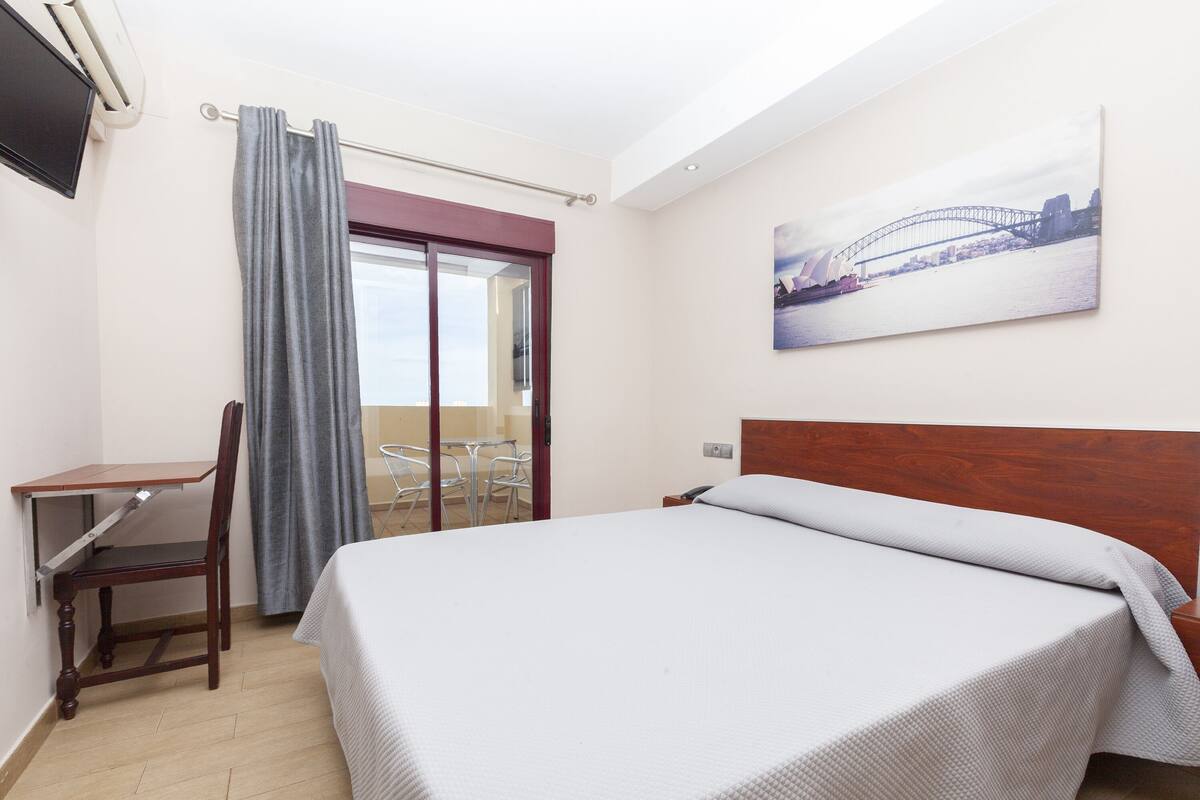 A hotel room with a bed, a desk, a chair, a window with curtains, and a painting of a bridge.