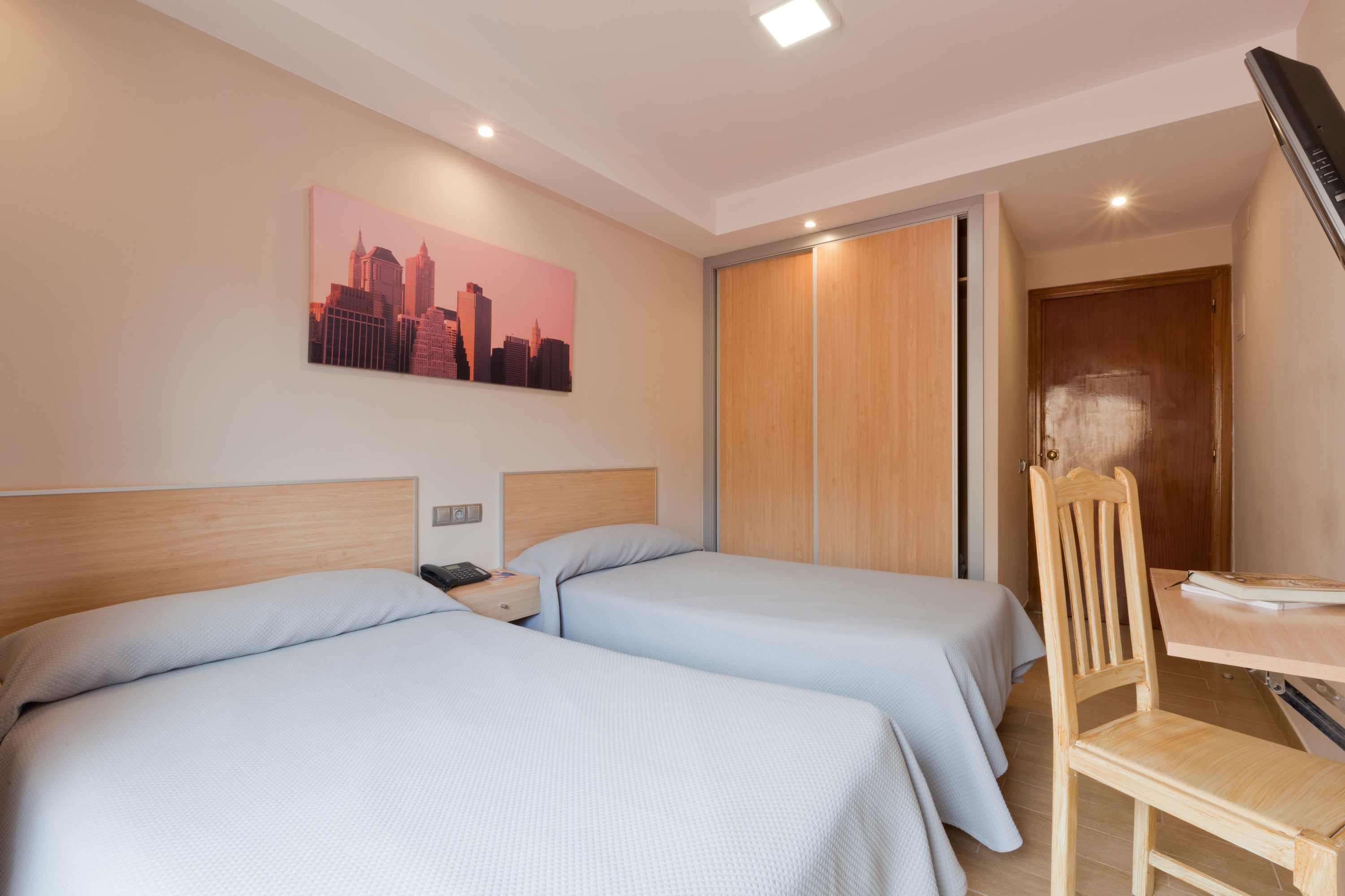 A hotel room with two beds, a wooden chair, a desk, a cityscape painting, and a sliding door.
