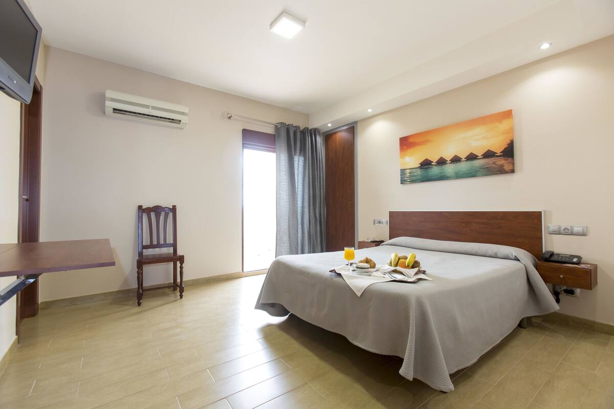 A hotel room with a bed, a desk, a chair, an air conditioner, a painting, and a window with curtains.